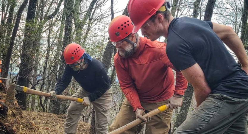 People wearing helmets use tools to work on a trail in a wooded area. 